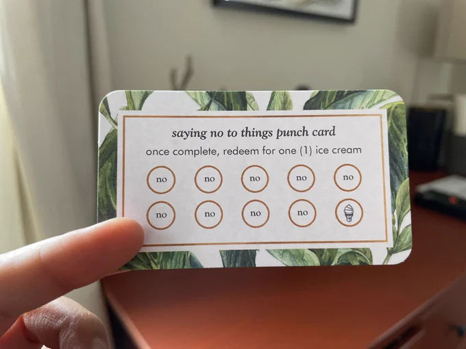A business card with rounded corners that has a watercolor botanical background. In the foreground: “saying no to things punch card. Once complete, redeem for one (1) ice cream,” followed by 9 circles that say “no” inside and one with a drawing of an ice cream