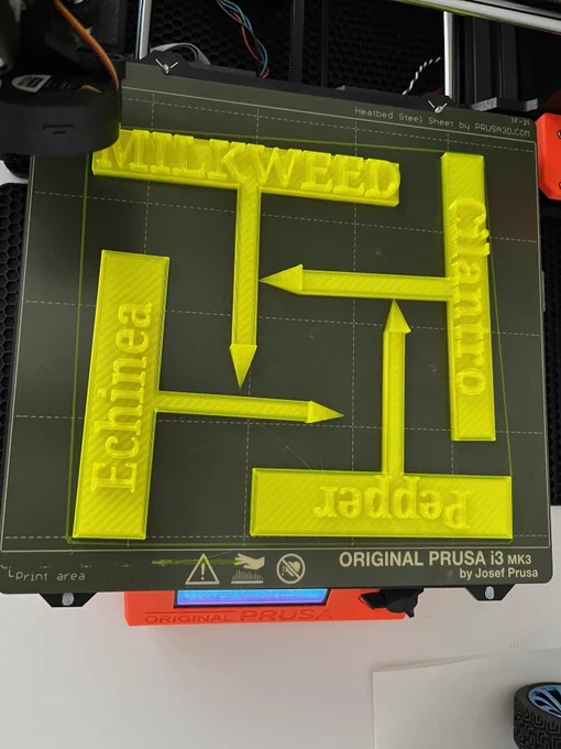 A 3D printer bed shows four garden spike signs, for pepper, cilantro, echinea and milkweed. 

