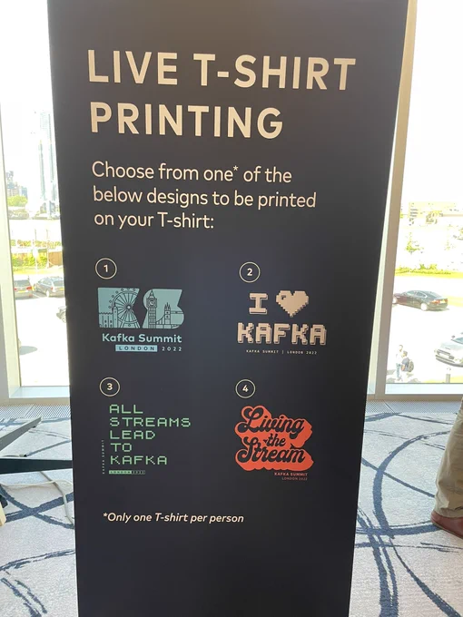 A standing poster at the Kafka summit conference in front of glass windows. The poster reads:

„Live T-Shirt printing

Choose from one* of the below designs to be printed on your T-Shirt:

[four different designs shown with the Kafka Summit logo, a „I love Kafka“ slogan, „All streams lead to Kafka“ and „Living the Stream“]


*Only one T-Shirt per person“
