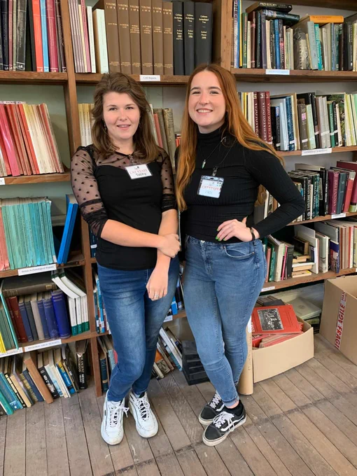Tabitha and Vikki stand in front of the C.A.T. bookshelves. They are both wearing blue jeans and black shirts.