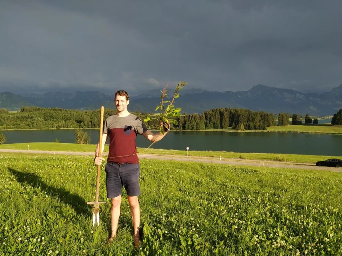 Photo of a man with a rake standing in a field. Behind him, a lake and a forest.
