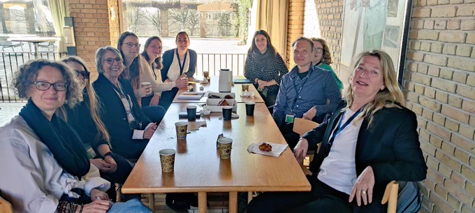 A group of people sitting at a big table indoors with bright morning light behind them and lots of coffee and pastries on the table.