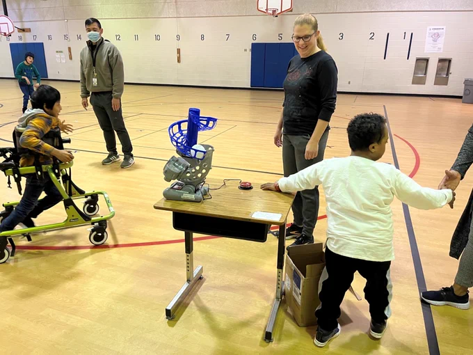 student standing next to adapted pitching machine place don table. ready to press red switch to activate machine to throw the ball.
