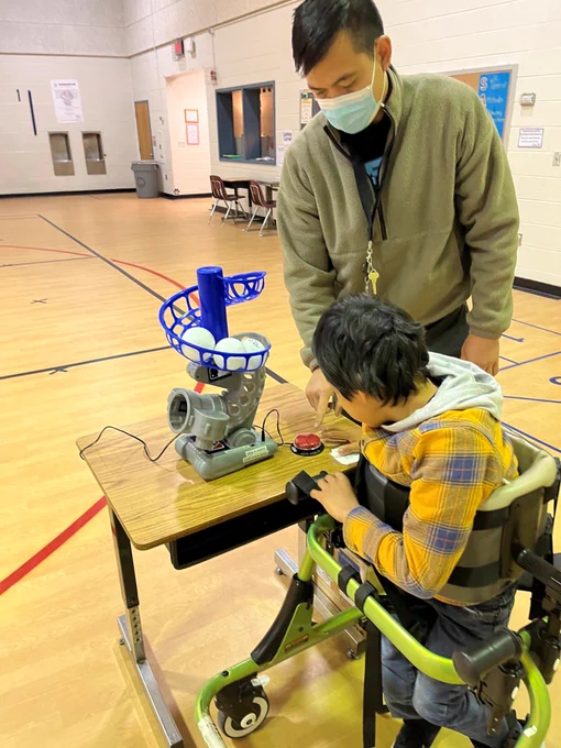 picture of student in wheelchair with help of instructional assistant pressing a red switch to activate pitching machine.