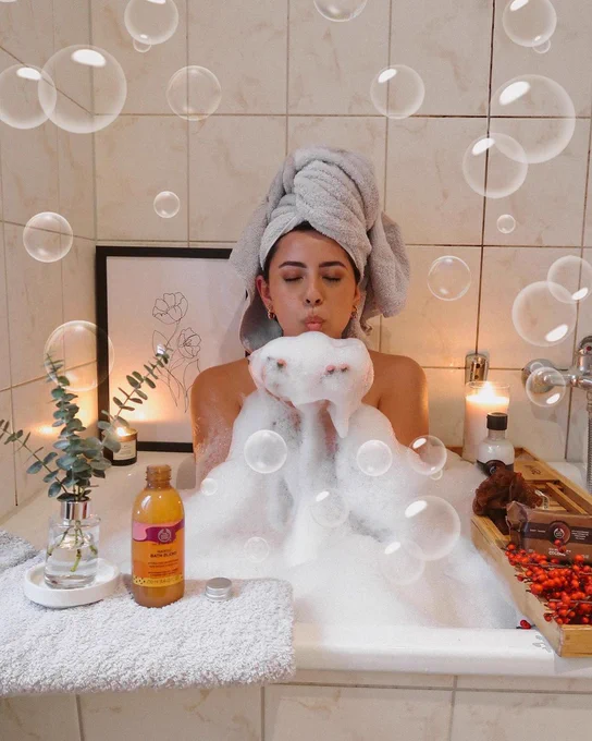 Face on image of Andy Chajón in a soapy bathtub, wearing a grey towel on her head. She's holding some suds in her hands and blowing toward the camera. Theres a small glass vase with some eucalyptus and a Mango Bath Blend from The Body Shop at the front of the tub, and items from The Body Shop's Coconut range to the right.