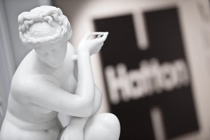 A marble sculpture of a woman in pose. In the background on the wall is the logo for the Hatton Gallery, Newcastle.