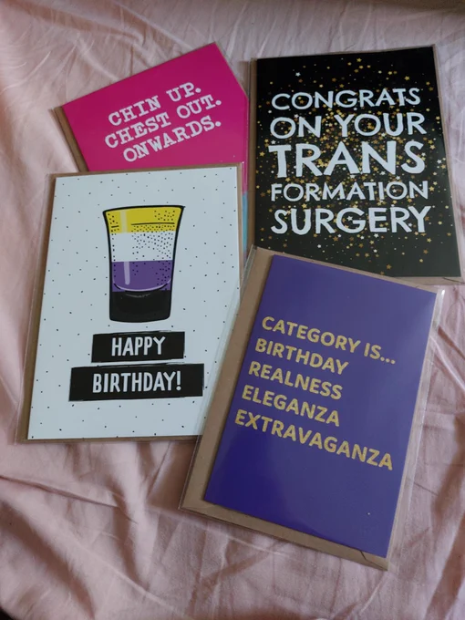 A selection of 4 greetings cards. Clockwise from top left is a bright pink card with white text reading “Chin up. Chest out. Onwards.”  Next is a black and gold card with white text reading “Congrats on your TRANS formation surgery”. Bottom right is a purple card with gold text reading “Category is… birthday realness eleganza extravaganza”. The last is a white card with non-binary flag colours on it reading “happy birthday!“. 