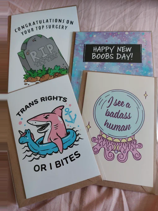 A selection of 4 greetings cards. Clockwise from top left is a tombstone reading RIP titties and “congratulations on your top surgery” on a white background. The next reads “happy new boobs day!”. Bottom right is a crystal ball on a cream background with text saying “I see a badass human”. Next is a pink shark riding a blue unicorn ring floatie, the text reads “trans rights or I bites”. 