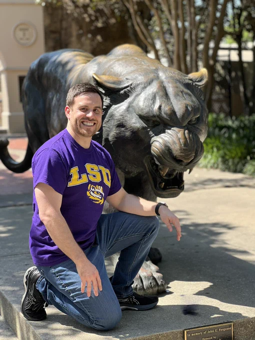 Smiling white man in a purple LSU t shirt kneeling in front of bronze tiger statue