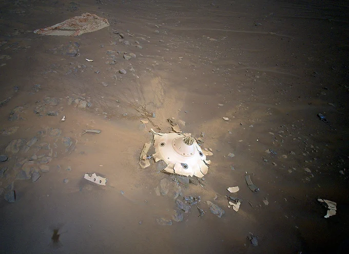 Camera view of the rover's backshell having impacted the surface of Mars. It looks like a hat with a flattened, shattered brim. The ground is an odixided burnt orange and brown, with some shades of gray. It's flat and relatively rocky. The backshell is a white color and looks metallic. Small bits of debris surround it.