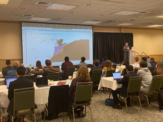 Jong Lee gives a talk at Colorado State University in a large conference room full of people. The screen shows a visualization of the Galveston coastline.