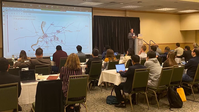 Jong Lee gives a talk at Colorado State University in a large conference room full of people. The screen shows an interactive visualization of a community with key locations highlighted.