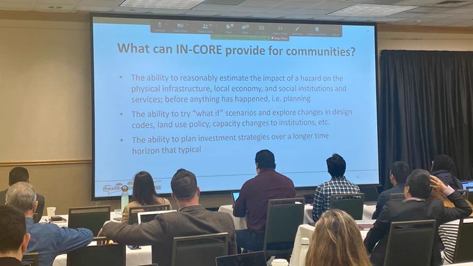 Sara Hamideh presents virtually about IN-CORE community partnerships to a large conference room full of people. The screen highlights what IN-CORE can provide for communities.