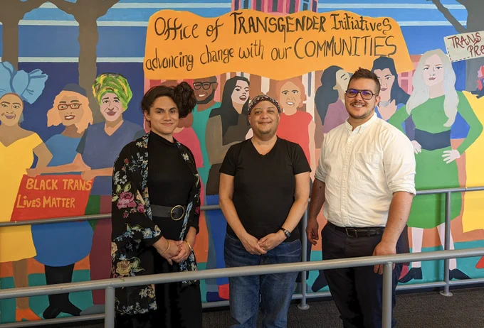 Photo of Shane, Pau, and Pax standing in front of a mural featuring trans people, a sign reading "Black Trans Lives Matter", and a banner reading "Office of Transgender Initiatives - advancing change with our communities". 
