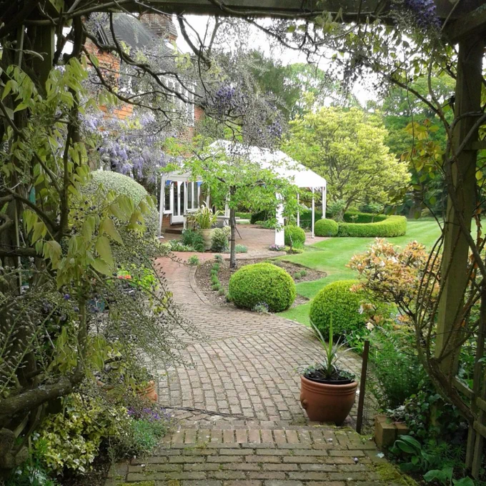 A white gazebo in the center of the frame, pictured through a wooden arch with small trees and bushes. 