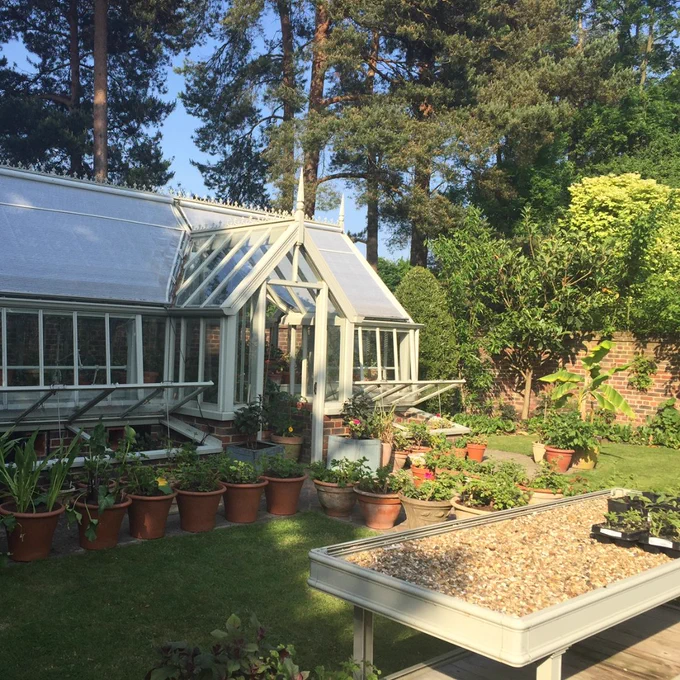 A white greenhouse in a large garden with a range of potted plants placed in front of it.