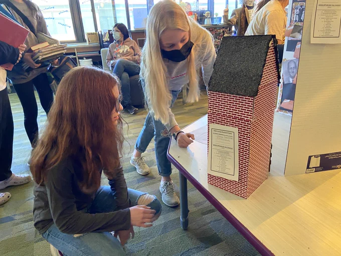 Two students stoop down to examine a diorama shaped like a house.