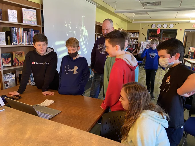 A group of students and a teacher gather around a table, looking at a laptop.