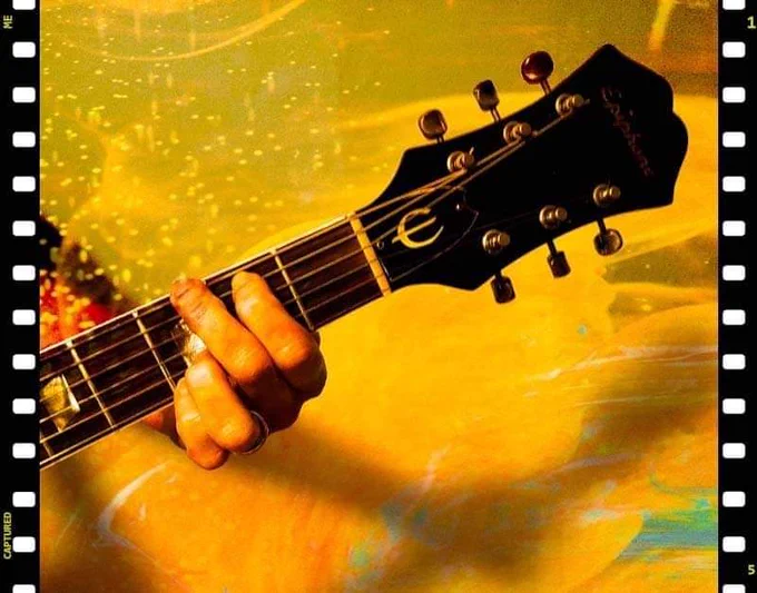 Six string guitar headstock and upper fretboard with a hand playing a chord. Yellow background. 