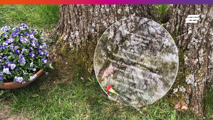 Foto: Auf einer grünen Wiese sind links eine Blumenschüssel mit lila Blumen zu sehen. Daneben ragen zwei dicke Bäume aus der Wiese, von denen nur die Ansätze zu sehen sind. Am Fuß der Bäume steht eine kreisrunde Glasscheibe auf der folgender Text eingraviert ist: "Hier wird im Herbst 2022 eine Geenkkugel aus Ton liegen: In Gedenken an alle lesbischen Frauen und Mädchen im Frauen-KZ Ravensbrück und Uckermark. Sie wurden verfolgt, inhaftiert, auch ermordet. Ihr seid nicht vergessen." Davor liegt eine einzelne rote Nelke.