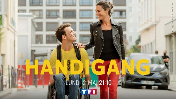 L'image est une publicité pour l e film Handigang. Au milieu de l'image se tiennent une femme et un jeune homme côte à côte. La femme est debout, une main posée sur l'épaule du jeune homme, assis dans son fauteuil roulant. Les deux protagonistes se regardent en souriant.