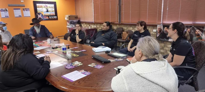A  group of people are gathered around a table chatting. There are AEC flyers placed on the table in front of the people. 