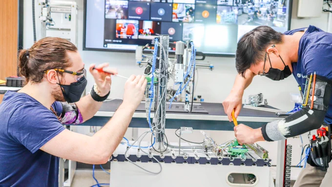Two students use handheld tools to work on a mechanical device in a lab.