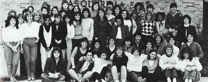 A large group of women stand and sit in front of Pasquerilla East Hall at the University of Notre Dame. Many have their arms around each other as they smile for the camera.