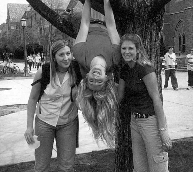 Two women stand near a tree smiling as they lean towards a third woman who is hanging upside down from the tree in between them.