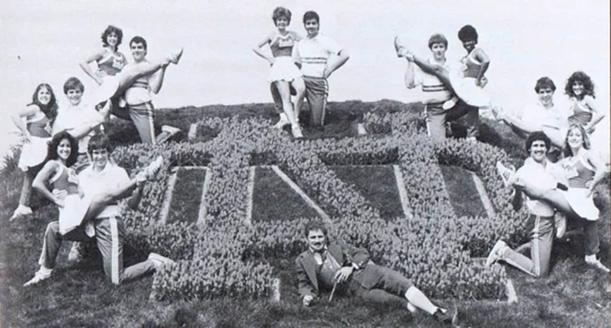 Seven female cheerleaders sit on a male cheerleading partner's knee with their leg extended surrounding an interlocking N and D Notre Dame logo. A man dressed as a leprechaun lounges at the bottom of the ND.