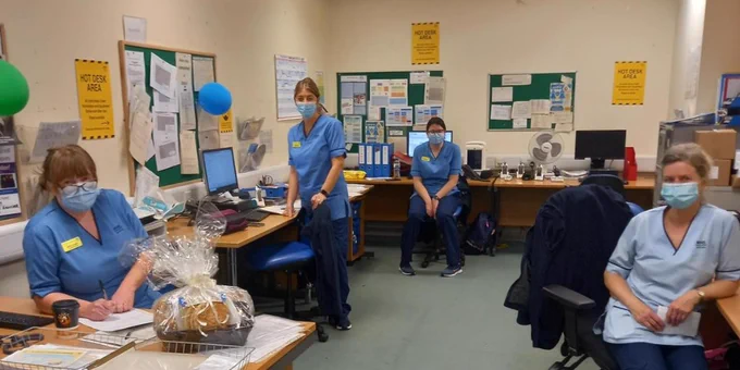 Four midwives, in a office with balloons. Three are sitting, one is standing, all face the camera.