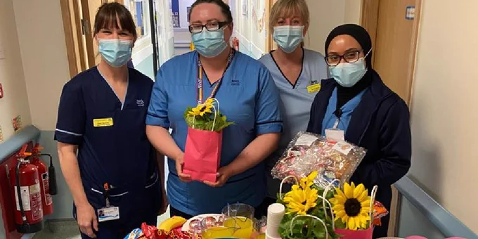 Four midwives, standing behind a tray decorated with flowers, food, and pink balloons. The second from left is holding a gift of flowers, the furthest-right midwife is holding a parcel of treats.