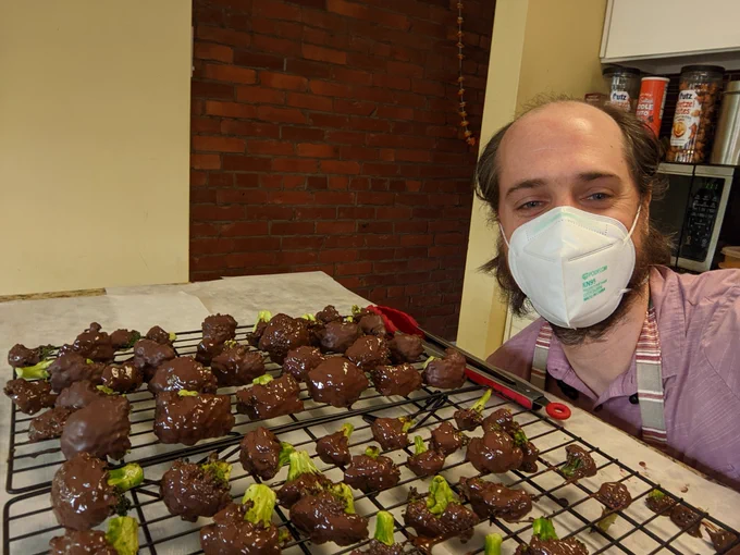 A man in an N95 mask crouching down next to trays of drying broccoli florets covered in chocolate.