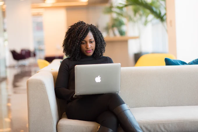 A dark-skinned woman sits on a white sofa while holding a laptop in her lap.