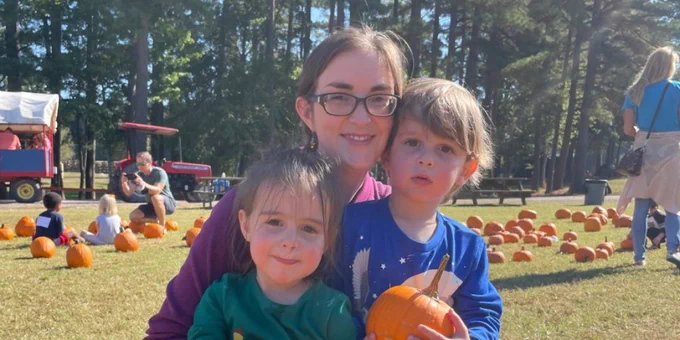 A women with dark hair and glasses wearing a purple shirt holds two small boys, one in a green shirt, the other in a blue shirt. The boys are holding small pumpkins. There are people picking pumpkins in the background.