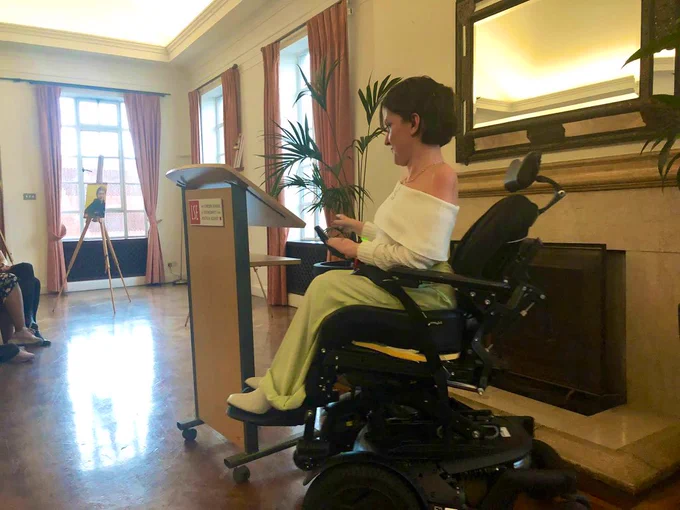 Anna, a white woman with short brown hair in a black power wheelchair, sits at the front of the room giving brief remarks after receiving her award.