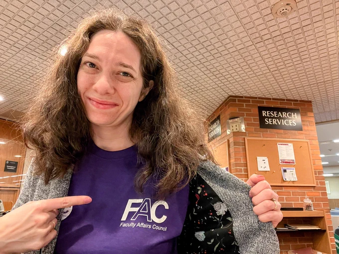 Smiling research services librarian sitting at the Research Services desk pointing to her union logo on the front of her purple shirt worn beneath a grey blazer.