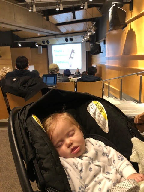 A small boy naps in a baby carrier in a lecture hall. In the background, several people are seated and listening to a scientific talk.
