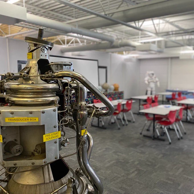 Photo of the Exploration Station classroom with rows of tables and tables, an astronaut suit in the background, and a rocket engine assembly in the foreground