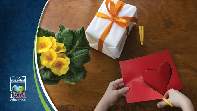 Bird's eye view of a brown table with yellow flower, wrapped gift and child's hands cutting a heart out of red paper. Green swoop across upper right corner with AMDSB logo.