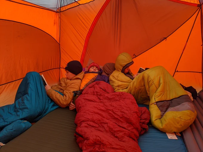A woman and two boys huddle together in orange sleeping bags in an orange tent. They are wearing warm knitted hats, and the boys are reading books. The woman between them is asleep.