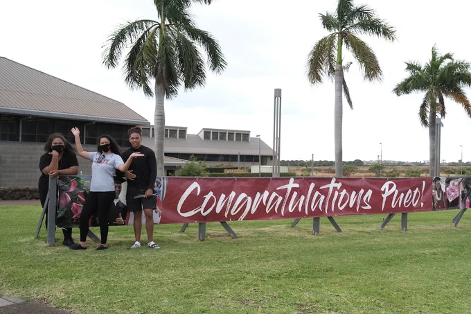Three people outdoors and standing by a banner that says, "Congratulations Pueo!"