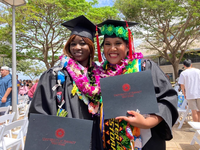 Two graduates wearing caps and gowns, and holding diploma covers.