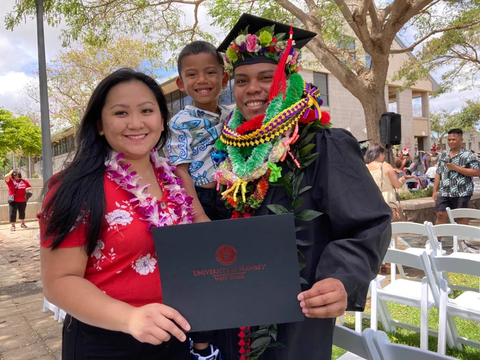 A graduate carrying a young boy, holding a diploma cover, and standing next to a woman.