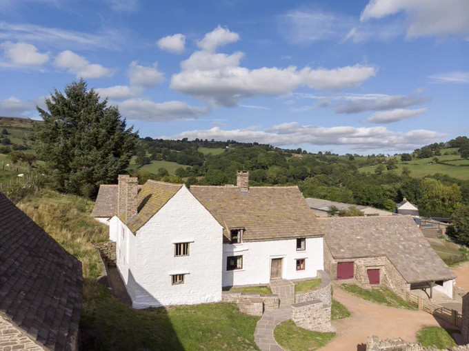 A white 15th century Welsh farmhouse standing amidst green rolling hills and a blue sky with white fluffy clouds.