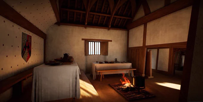 An interior shot of the high end of a hall in the 15th century farmhouse. A table with a white cloth is laden with food and there is a fire lit in the centre of the room. The ceilings are exposed wooden beams.
