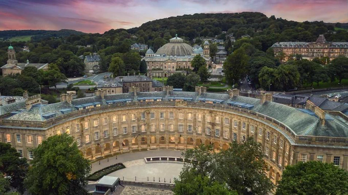 The exterior of Buxton Crescent, with the landscape of Buxton, trees and sunset behind it.