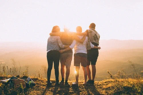 four people stand in silhouette on a hill overlooking a vista at sunset, with arms wrapped around each other's backs in support.