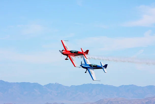 Two fighter planes fly parallel to each other. The top one is red, and the other is blue. The red plane emits a cloud of smoke.