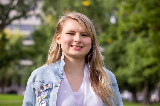 A young person with long blonde hair smiling at the camera. They are in a park. They are wearing a denim jacket with pride badges on it.